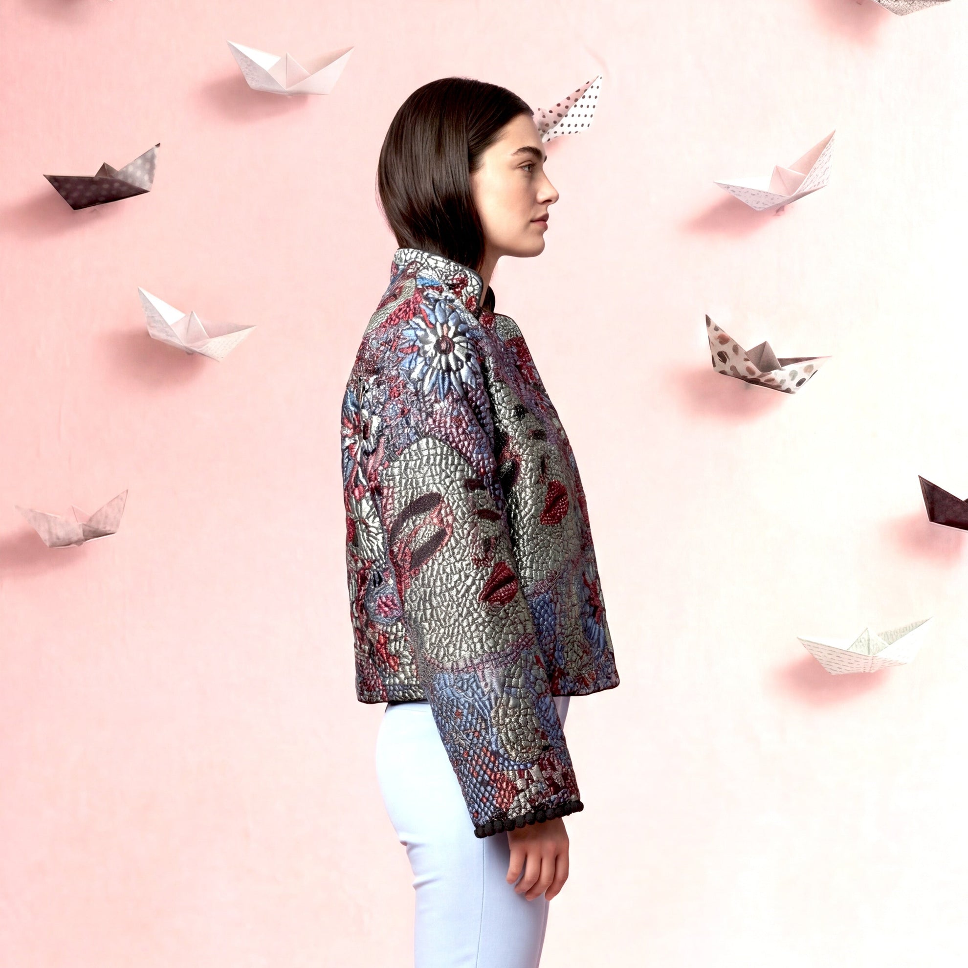 Woman wearing a patterned jacket and light blue pants against a pink background with paper boats.