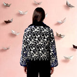Person wearing a patterned blouse and black pants standing against a pink background with paper boats.