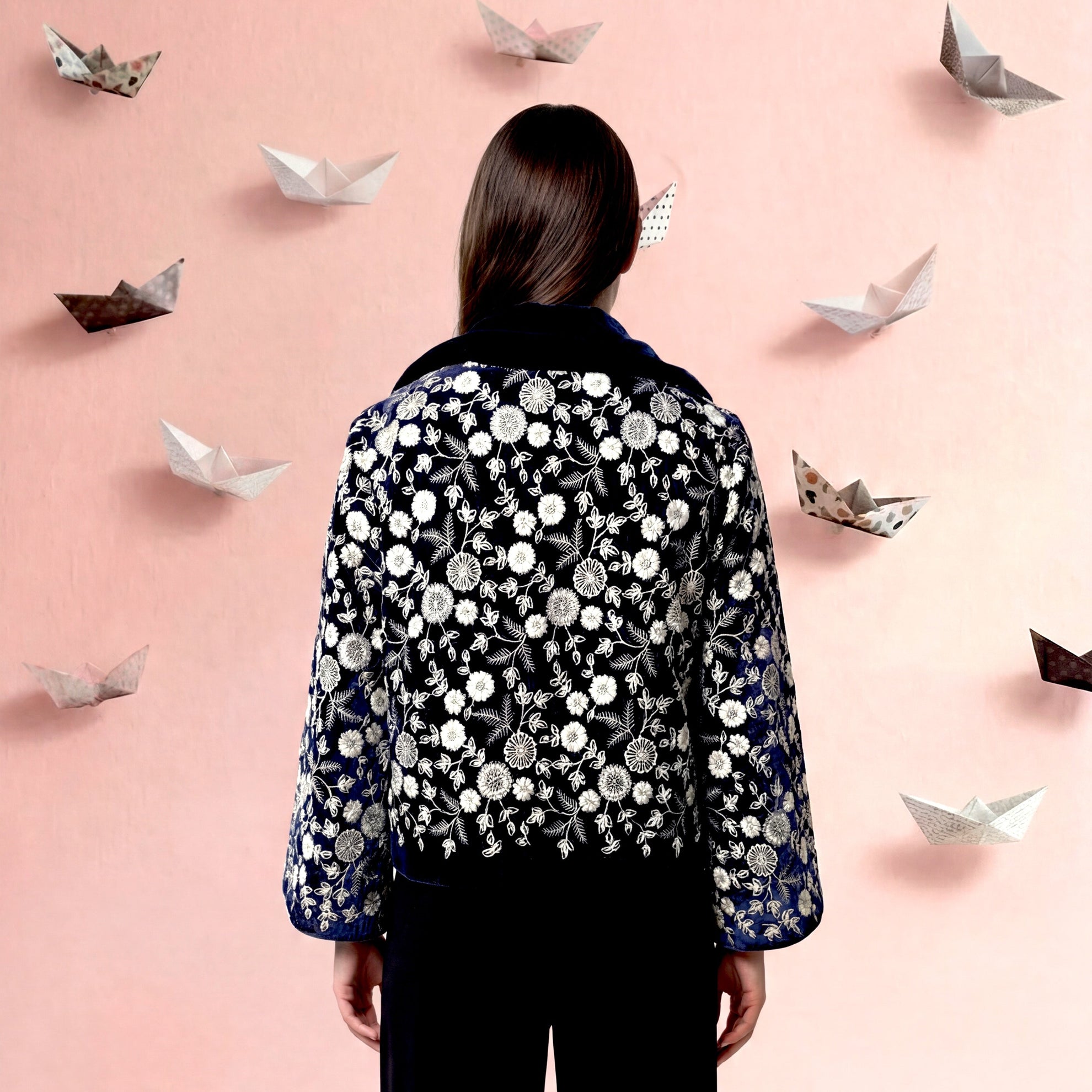Person wearing a patterned blouse and black pants standing against a pink background with paper boats.