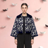 Woman wearing a floral jacket and black pants with paper swans and boats on a pink background