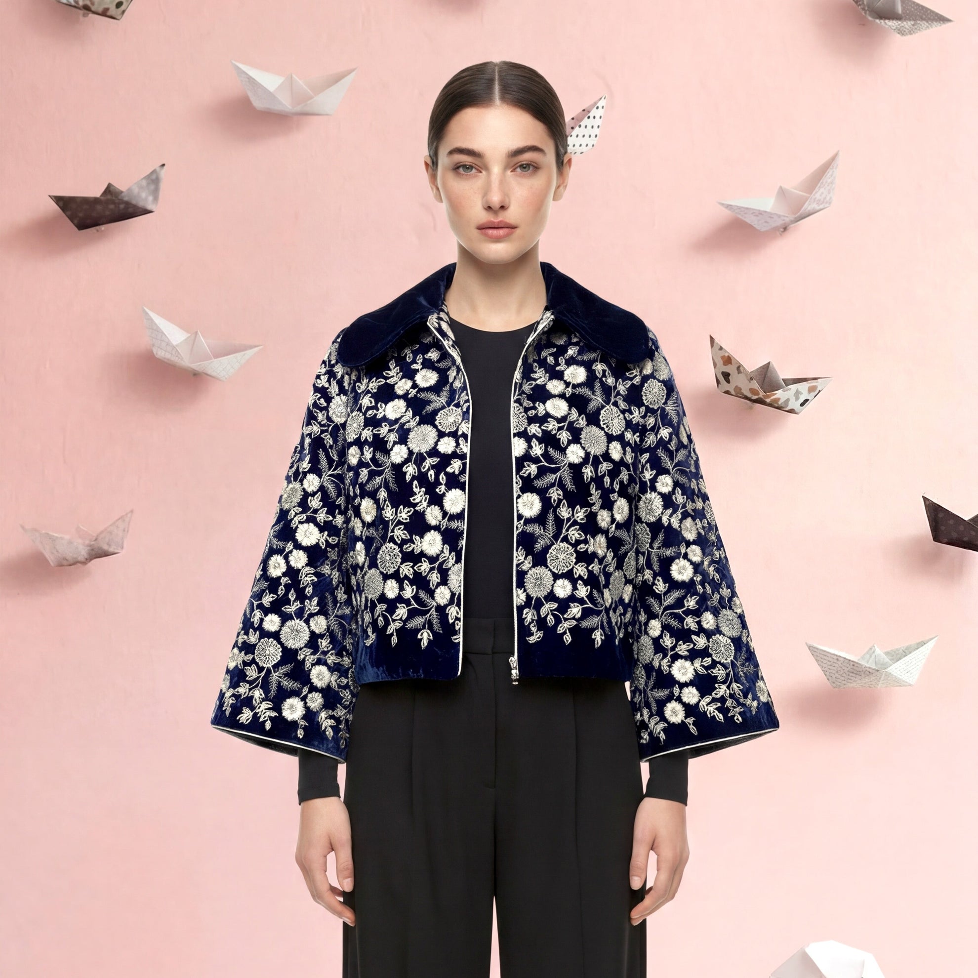 Woman wearing a floral jacket and black pants with paper swans and boats on a pink background